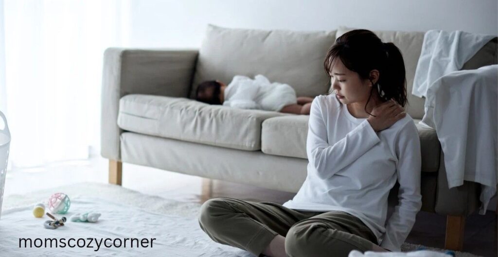 A tired mother sitting on the floor leaning against a couch with her head turned away while a baby sleeps in the background.