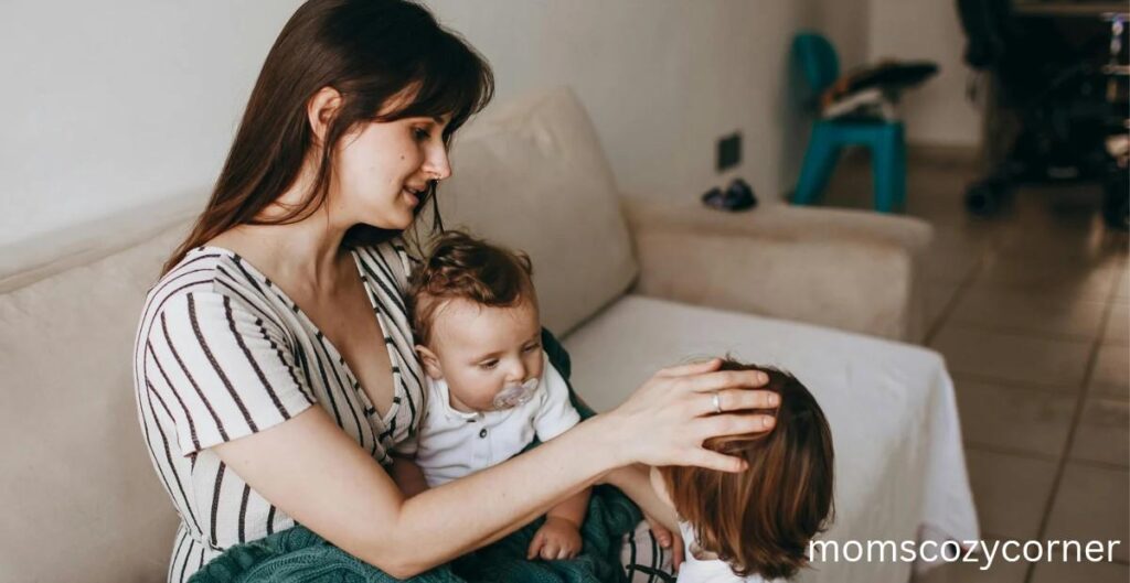 A mother sitting on a couch smiling gently and touching her child's head while holding a baby.