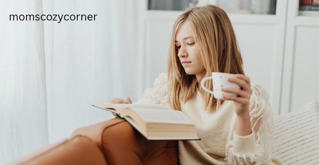 A young woman sitting comfortably while reading a book and holding a white coffee mug.